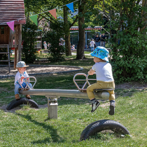 Zwei kleine Kinder spielen auf einer Wippe im Außenbereich der BEST-Sabel Kita Kaulsdorf, umgeben von Bäumen, Spielgeräten und bunten Wimpeln an einem sonnigen Tag.