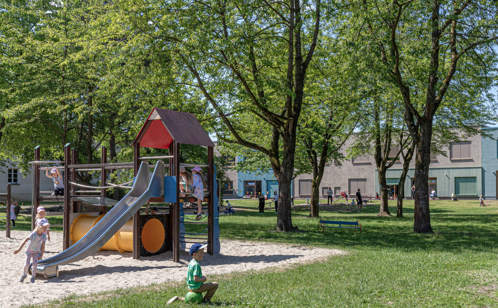 Kinder spielen an einem sonnigen Tag im Garten der Kita Kaulsdorf. Im Vordergrund ist eine Rutsche und ein Kletterturm zu sehen.